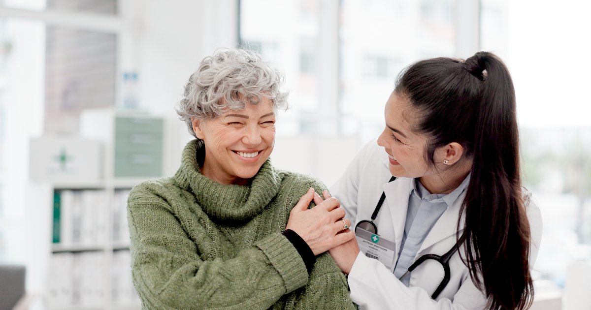 A smiling older woman in a green sweater holds hands with a doctor in a white coat in a bright clinic.